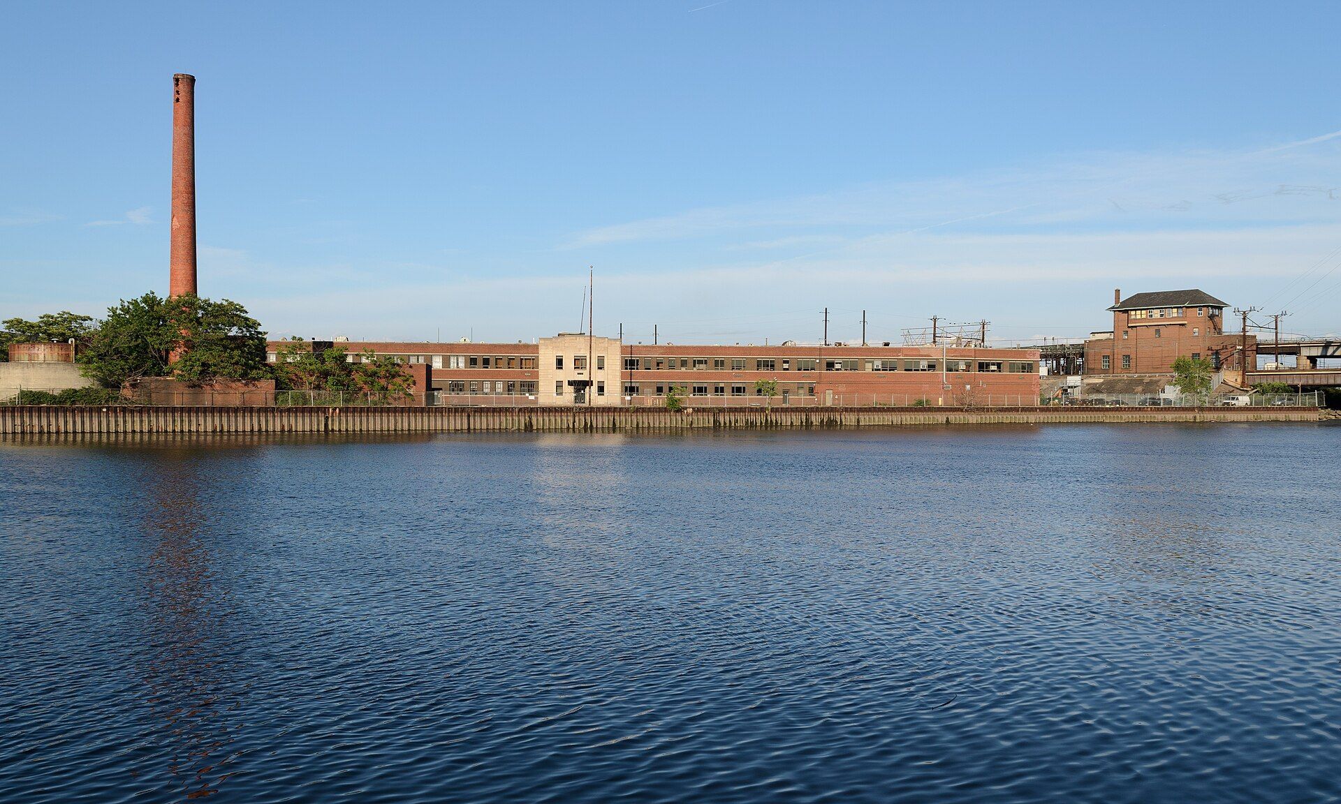 A long, low brick industrial building with a tall smokestack on the bank of the Passaic River in Harrison, New Jersey.