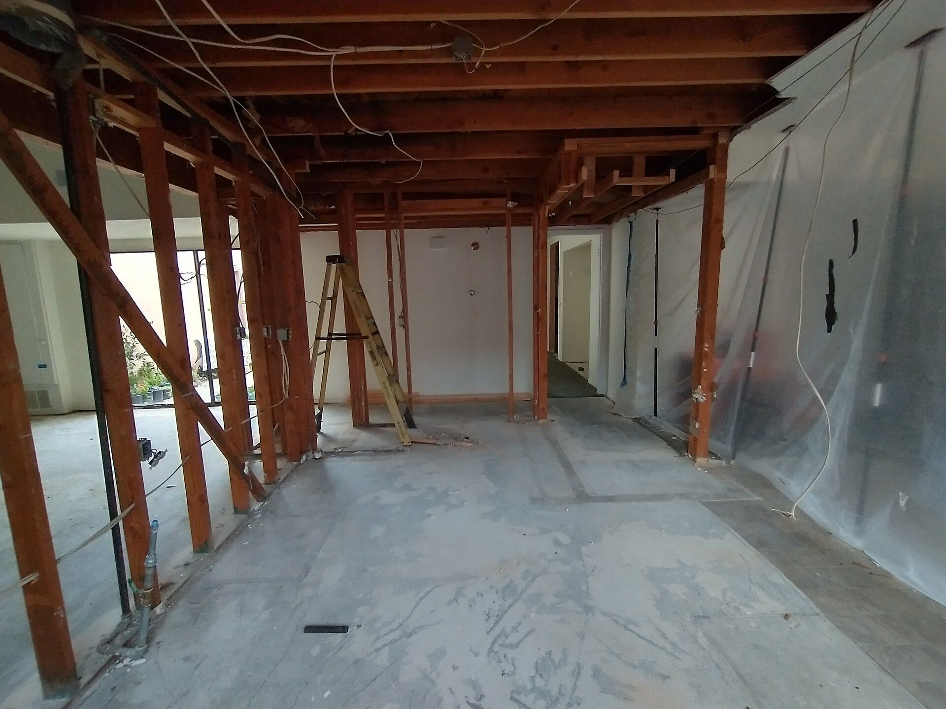 A gutted interior with exposed wood stud walls and ceiling joists, loose electrical wiring and a bare concrete floor, with plastic sheeting hung along one side.