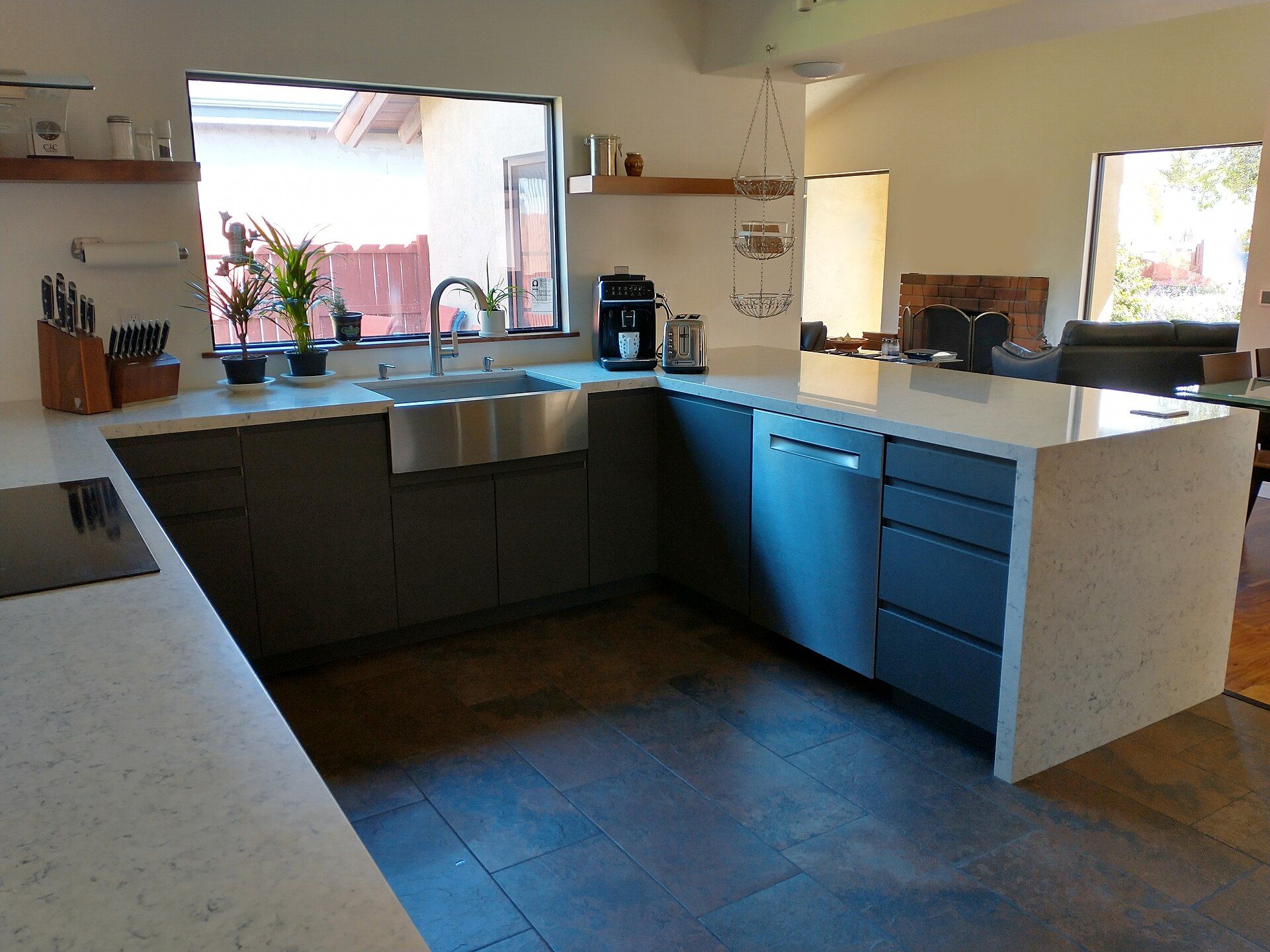 A renovated kitchen with gray flat-panel cabinets, white quartz countertops, a stainless sink under a large window, a built-in dishwasher and a cooktop.