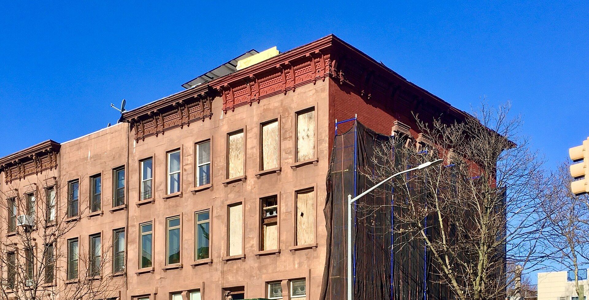 A row of brownstone row houses on a Brooklyn corner, the end building wrapped in black netting and scaffolding, its windows boarded for renovation.
